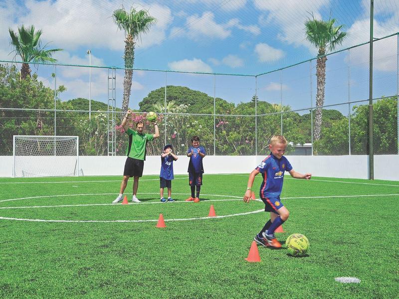 Kinder trainieren Fußball auf grünem Kunstrasen mit Hütchen, Trainer beobachtet, sonniger Tag.