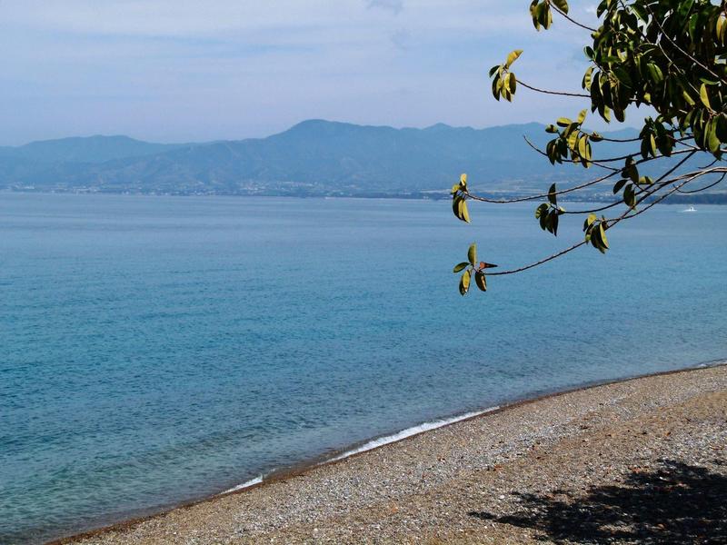 Klares blaues Wasser an Kiesstrand mit Bergkette im Hintergrund und Blättern im Vordergrund.