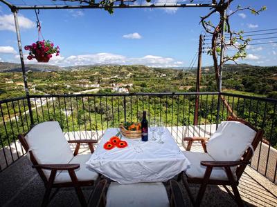 Balkon mit Tisch, zwei Stühlen, Weinflasche, Glas und Obst vor grüner Landschaft.