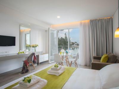 Bright hotel room with double bed, seating area, dining table, and balcony view of the sea.