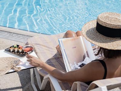 Person relaxing by pool wearing sunhat, reading magazine and drinking cocktail.