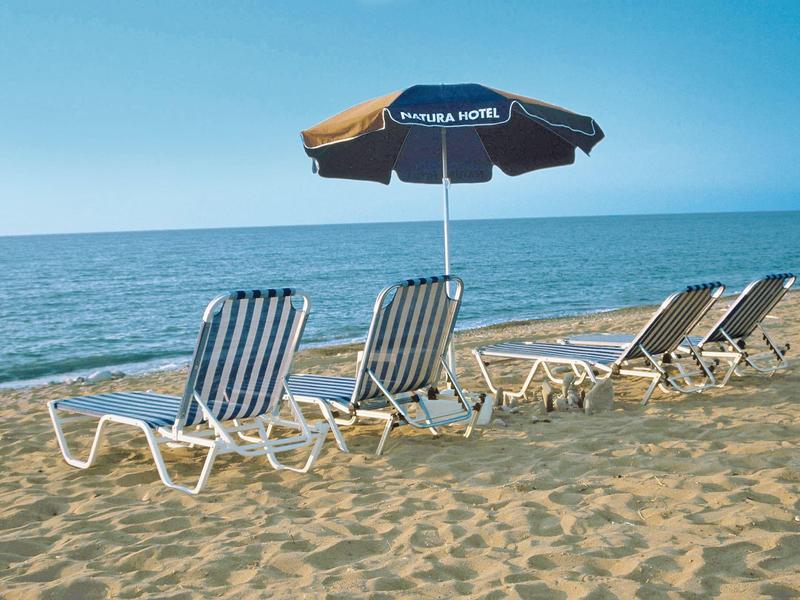 Vier Sonnenliegen und ein schwarzer Sonnenschirm stehen auf hellem Sand am ruhigen blauen Meer.