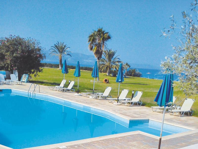 Ein klarer Pool mit weißen Liegestühlen und blauen Sonnenschirmen, grüner Rasen und Meer im Hintergrund.