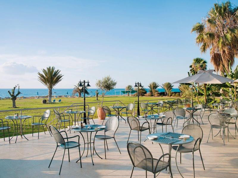Leerer Außenbereich mit Metalltischen und -stühlen, Palmen, Rasen und Blick aufs Meer.
