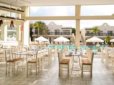 Zona pranzo luminosa con tavoli e sedie in legno, vista sulla piscina e ombrelloni