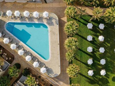 Ein blauer trapezförmiger Pool mit weißen Sonnenschirmen daneben auf grünem Rasen und Wegen.