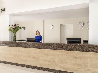 Recepcja hotelowa z uśmiechniętym pracownikiem i bukietem kwiatów na ladzie.