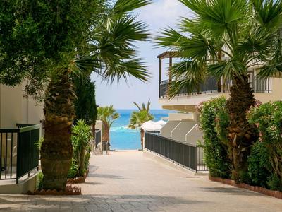 Sentiero tra edifici con palme che conduce a una spiaggia e al mare blu sullo sfondo.