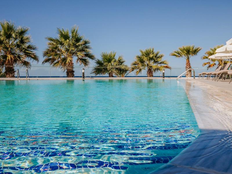Großer Pool mit klarem blauem Wasser, Palmen am Rand und Liegestühlen auf der Terrasse darunter.