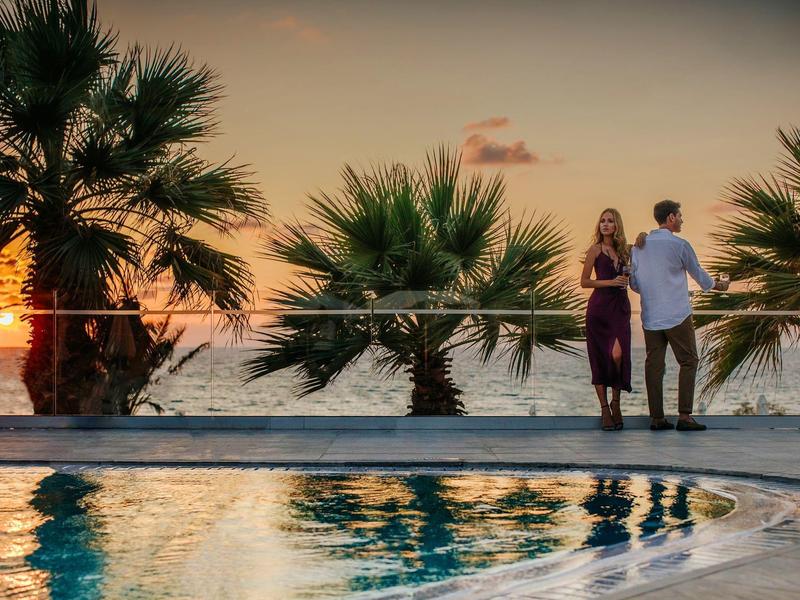 Ein Paar steht am Poolrand bei Sonnenuntergang, Palmen und Meer im Hintergrund.