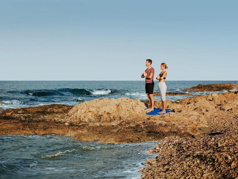 Zwei Personen stehen auf felsiger Küste mit Blick aufs Meer unter klarem Himmel.