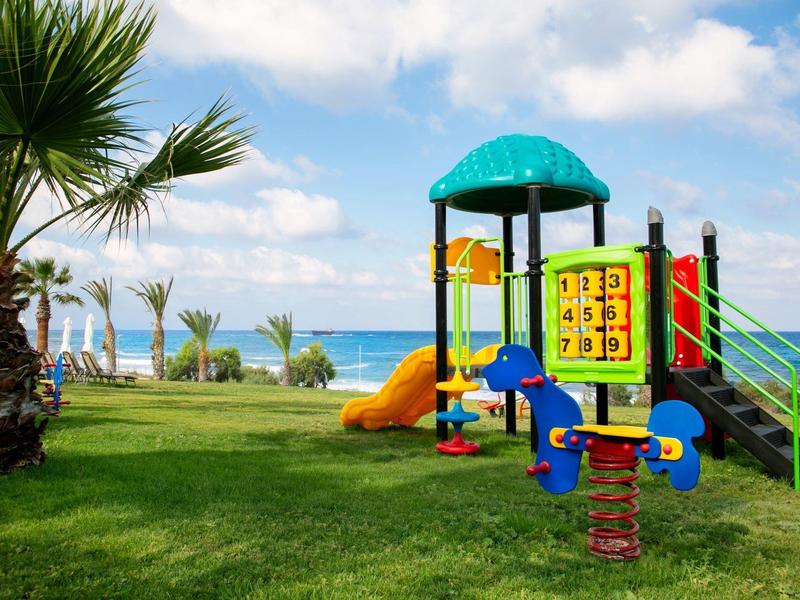 Bunter Spielplatz mit Rutsche auf grünem Gras nahe Palmen und blauem Meer unter blauem Himmel.