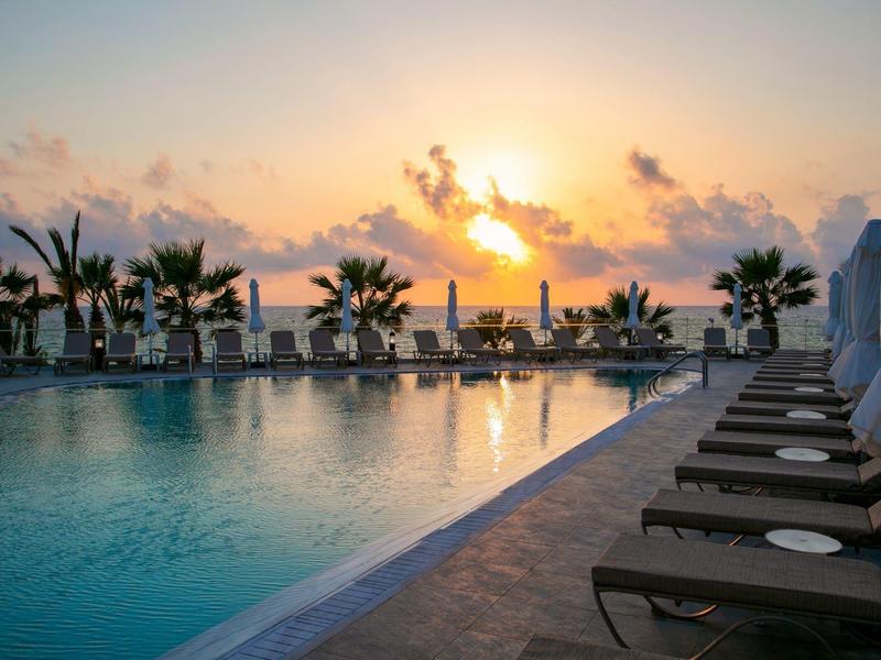 Schwimmbad mit Liegestühlen und Palmen bei Sonnenuntergang am Meer.