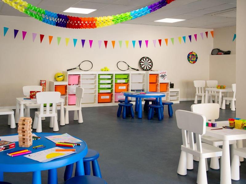 Bunte und helle Kinderspielecke mit kleinen Tischen, Stühlen, Regalen und Deko-Wimpeln.