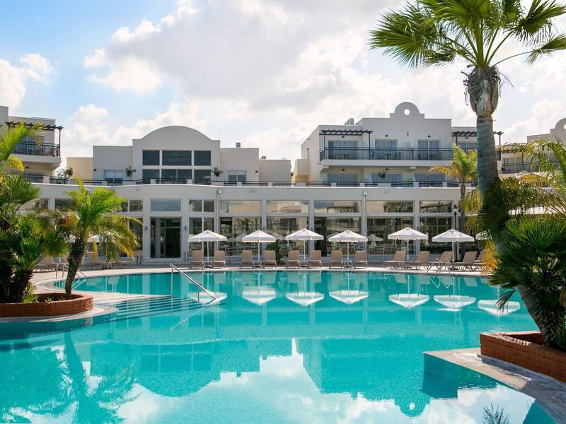 Großer Hotelpool mit Sonnenschirmen, Palmen und weißen Gebäuden im Hintergrund unter blauem Himmel.