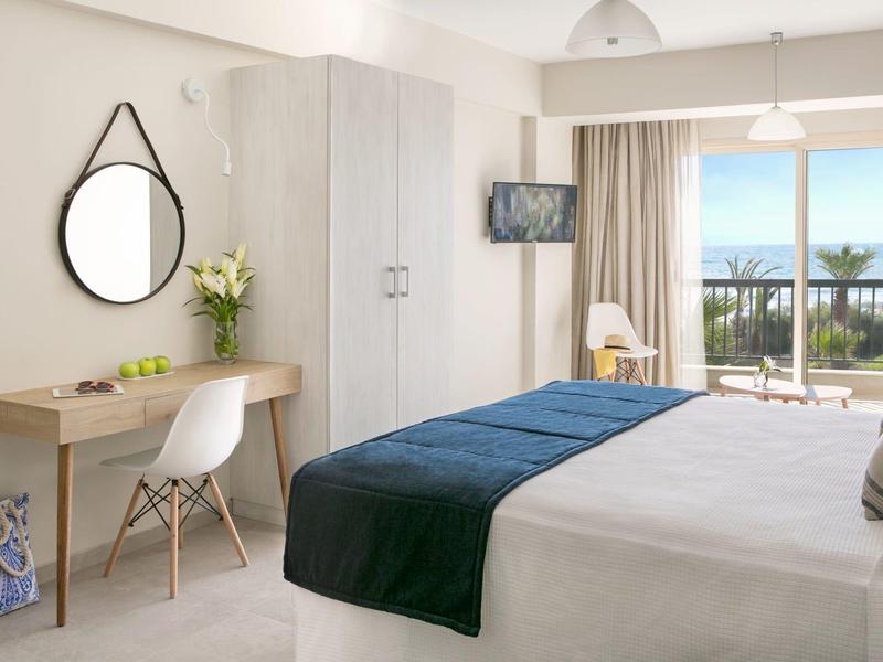 Helles Schlafzimmer mit weißem Bett, blauem Läufer, Schreibtisch, Rundspiegel und Balkon mit Meerblick.