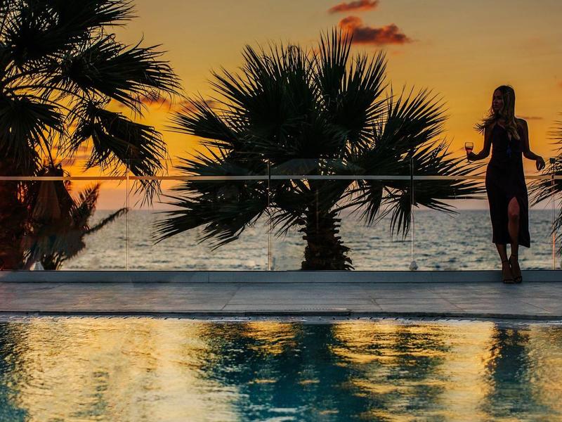 Frau steht am Poolrand bei Sonnenuntergang mit Palmen im Hintergrund, Wasser spiegelt Licht.