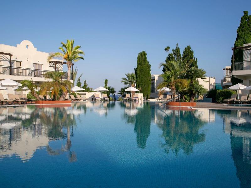 Großer, klarer Pool spiegelt Palmen, Gräser und weiße Hotelgebäude bei blauem Himmel wider.