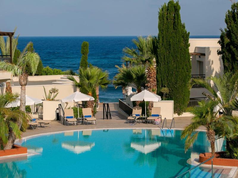 Pool mit blauer Wasserfläche, Palmen und Sonnenschirmen, dahinter Meer und Gebäude.