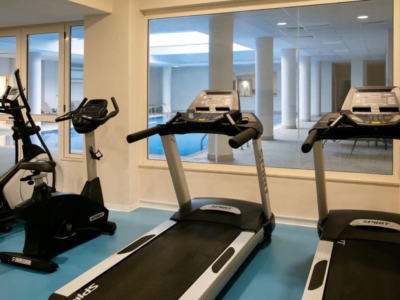 Fitnessraum mit Fahrradergometer und zwei Laufbändern vor großen Fenstern, heller Boden.