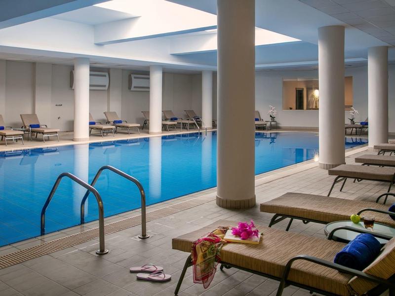 Innenpool mit blauer Wasseroberfläche, Liegestühlen, Säulen und Deckenfenstern für Tageslicht.