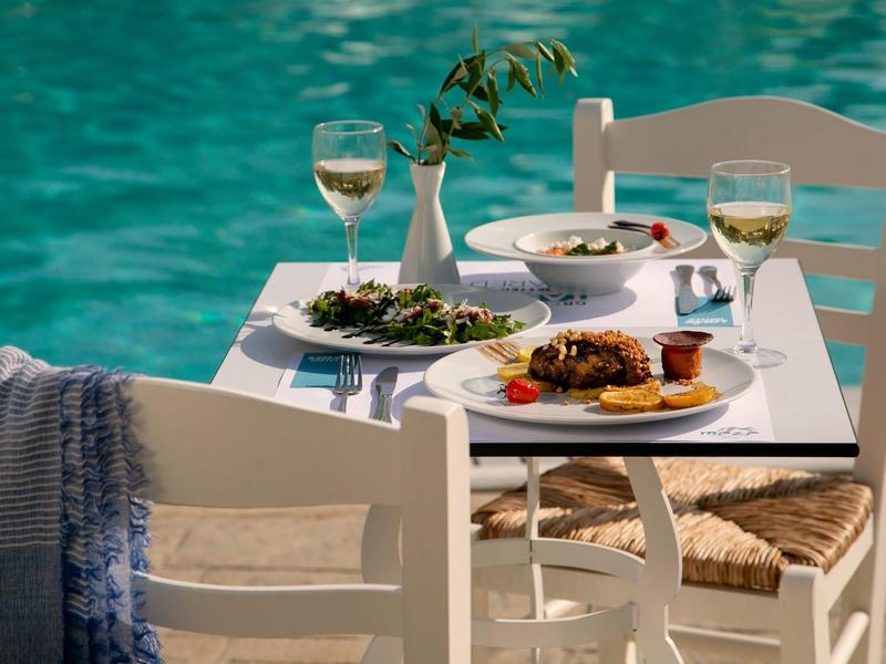 Weiß gedeckter Tisch mit Essen und Wein vor blauem Wasser, weiße Holzstühle, entspannte Atmosphäre.