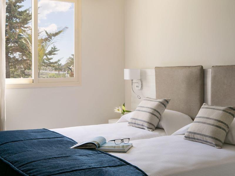 Helles Schlafzimmer mit großem Fenster, zwei Betten, grauen Kopfteilen und blauen Decken.