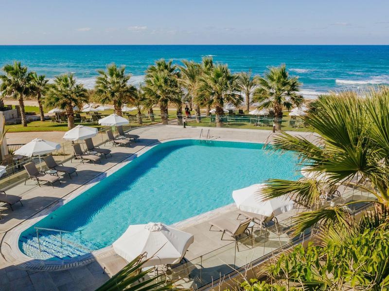 Rechteckiger Pool mit weißen Liegen und Schirmen, umgeben von Palmen und Meer im Hintergrund.