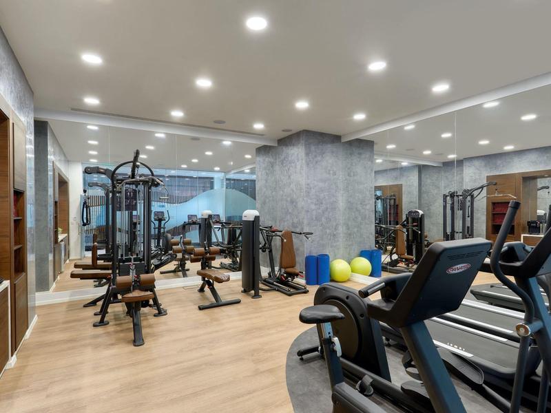 Moderner Fitnessraum mit Holzboden, Spiegelwänden, Trainingsgeräten und Deckenbeleuchtung.