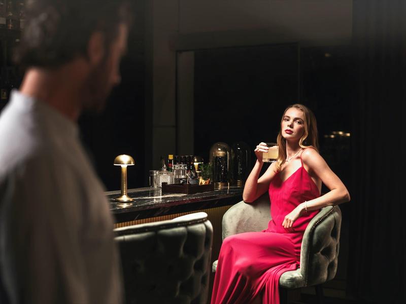 Frau im roten Kleid sitzt an elegantem Barhocker, Mann unscharf im Vordergrund, gedämpftes Licht.