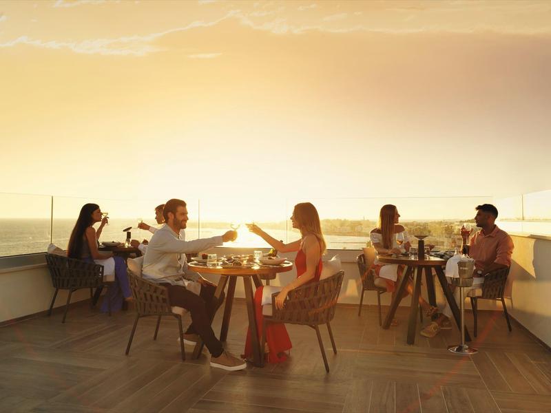 Dachterrasse mit Menschen, die bei Sonnenuntergang an Tischen sitzen und Getränke genießen.