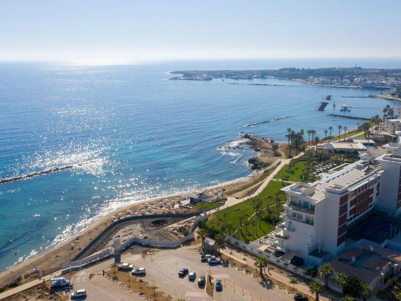 Küstenstadt mit blauer See, Sandstrand, Promenade, Gebäuden und grünen Flächen bei klarem Himmel.
