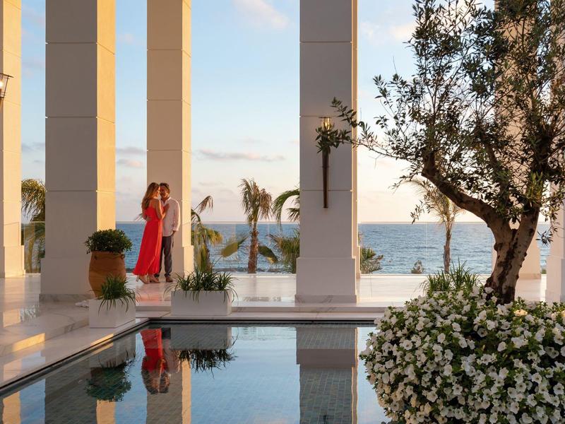 Zwei Personen am Meer vor weißen Säulen, neben reflektierendem Wasser und blühenden Pflanzen.