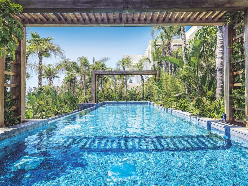 Rechteckiger Pool mit klarem blauem Wasser, umgeben von üppigen tropischen Pflanzen und Palmen.