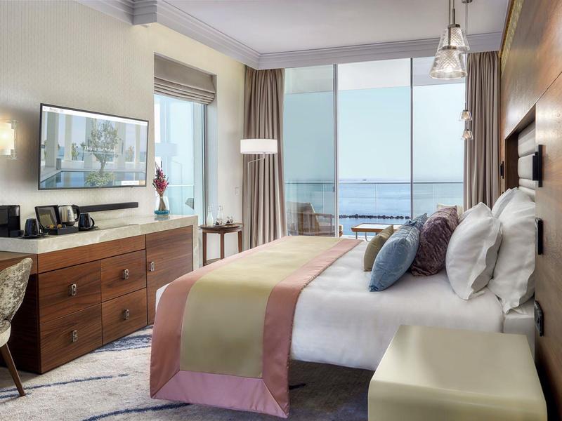 Modernes Hotelzimmer mit großem Bett, Holzmöbeln, Schreibtisch und Meerblick durch Panoramaglasfenster.