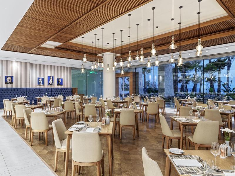 Modernes Restaurant mit Holzboden, weißen Stühlen, blauen Polstersitzen und großen Fenstern.