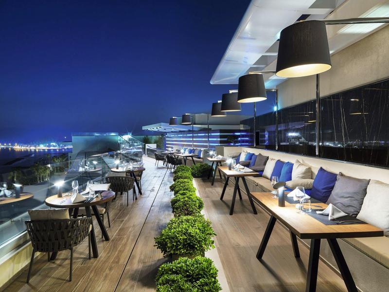 Moderne Dachterrasse mit gedeckten Tischen, Sitzbänken, Pflanzen und Stadtblick bei Nacht.