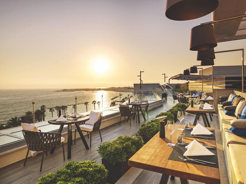 Terrassenrestaurant mit Tischen, Stühlen und Sonnenuntergang über dem Meer im Hintergrund.