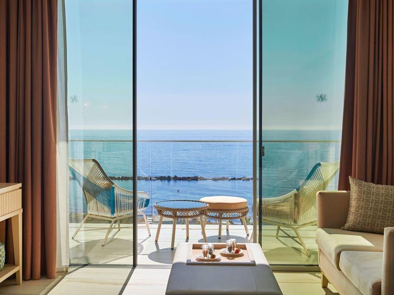 Moderner Wohnraum mit beige Sofa, Glasfront und Blick auf Terrasse und blaues Meer.