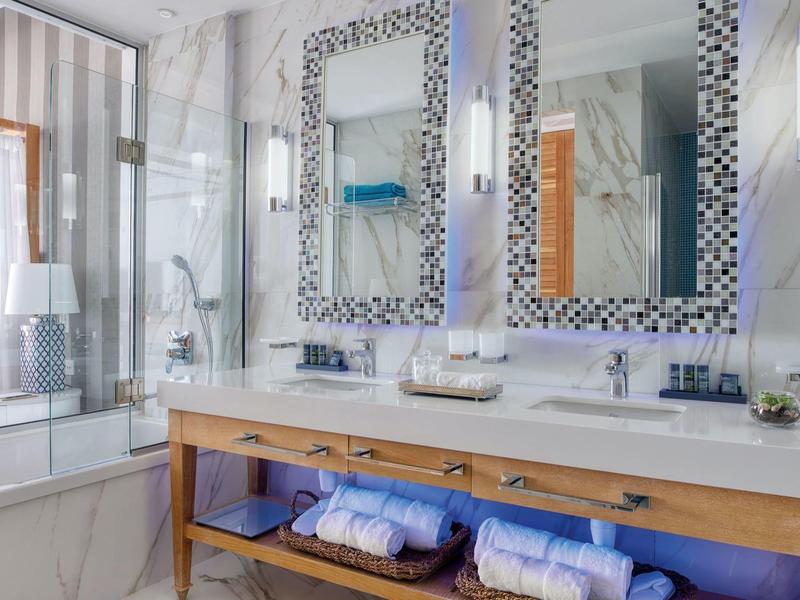 Helles Badezimmer mit Doppelwaschbecken, Mosaikspiegeln und Holzregal unter Marmorarbeitsplatte.