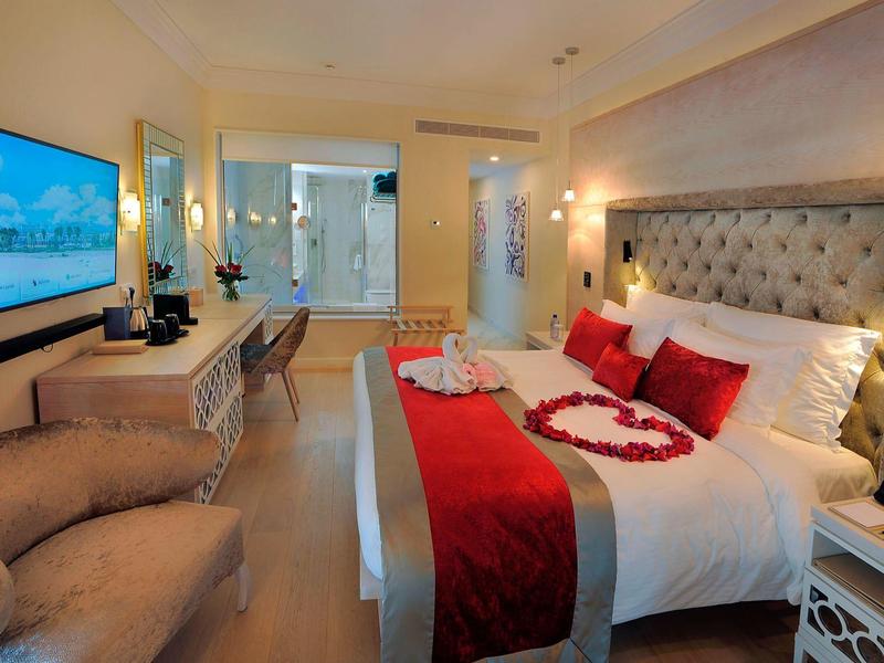 Modernes Hotelzimmer mit großem Bett, rote Dekokissen, Herz aus Rosenblättern und gemütlichem Sessel.