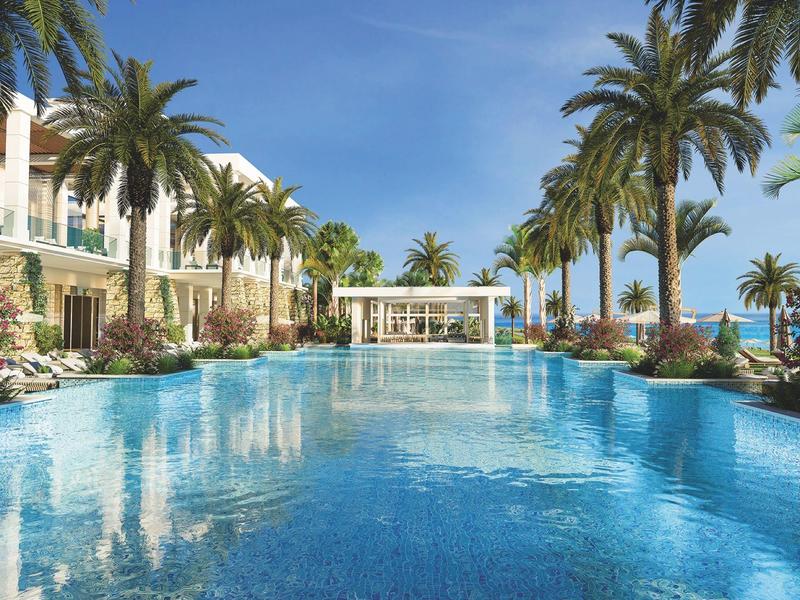Großer Pool mit klarem Wasser, umgeben von Palmen und weißen Gebäuden unter blauem Himmel.