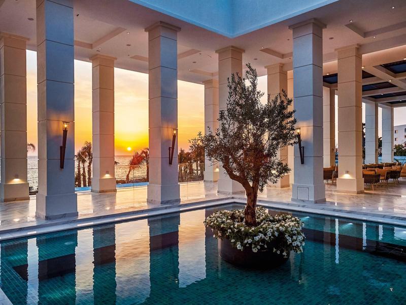 Moderner Innenhof mit Pool, großer Baum in der Mitte, Säulen und Sonnenuntergang am Meer.