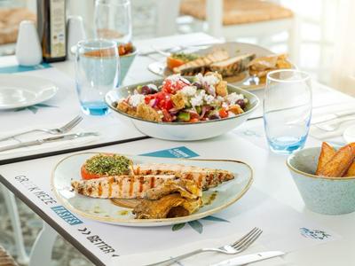 Tisch mit weißem Geschirr, gegrilltem Fisch, Salat, Brot und Gläsern in hellem Ambiente.