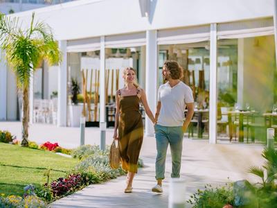 Couple marchant main dans la main devant un hôtel moderne à façade en verre par jour ensoleillé.