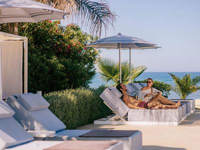 Zwei Personen entspannen auf weißen Liegen mit Sonnenschirm am Pool nahe Palmen und Meer.