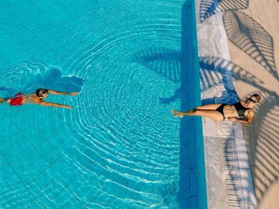 Donna che nuota in piscina limpida, uomo che prende il sole sul bordo con un asciugamano turchese.