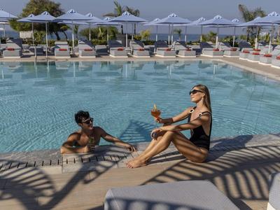 Piscina dell'hotel con due persone che si rilassano, circondate da ombrelloni e cielo azzurro.