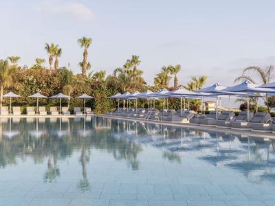 Grande piscina dell'hotel con sdraio e ombrelloni, circondata da palme sotto un cielo azzurro chiaro.