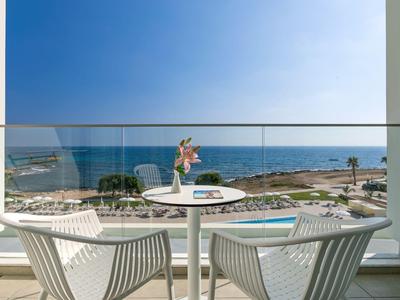 Balkon mit weißem Tisch, zwei Stühlen und Blick auf Meer, Pool und sonnigen Himmel.